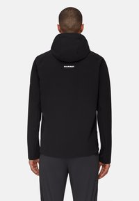 Personne portant une veste à capuche noire avec le logo "Mammut" dans le haut du dos, se tenant de dos sur un fond uni clair.
