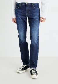 Dark blue denim jeans with a straight-leg design, five pockets, light fading, and stitched detailing. Paired with black high-top sneakers.