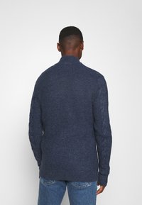 Man som bär en mörkblå stickad tröja med kabelmönster på ärmarna och blå jeans, stående vänd mot en neutral vit bakgrund.