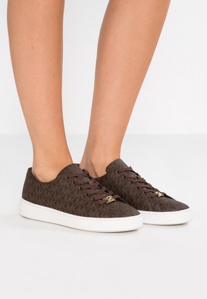 Personne portant des baskets basses marron avec motif monogramme, lacets marron et semelles blanches sur une surface blanche.