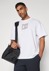 Mand iført hvid Under Armour t-shirt og mørke bukser med en sort skuldertaske, stående mod en ensfarvet lys baggrund.