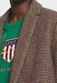 Bruin houndstooth blazer van textuurstof, met daaronder een groene trui met een embleem en gouden details op de borst.