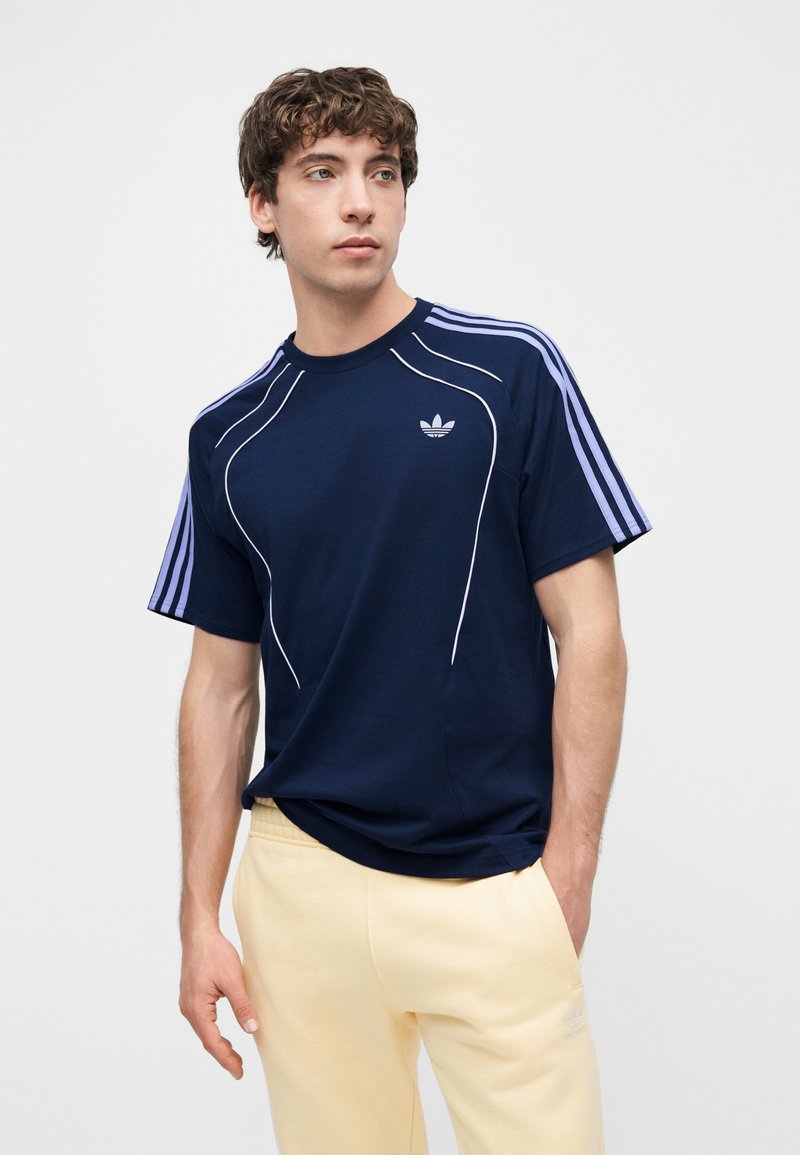 Young man wearing a navy blue Adidas shirt with light purple stripes and beige pants, standing with one hand in pocket.