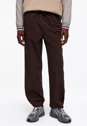 Homme portant un pantalon large marron foncé avec une taille coulissante et des baskets grises New Balance, debout devant un fond blanc.