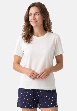 Vrouw met golvend bruin haar die een eenvoudig wit T-shirt met korte mouwen en marineblauwe korte broek met kleine witte patronen draagt, glimlacht en kijkt opzij.