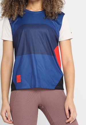 Frau trägt ein blaues, schwarzes, weißes und rotes kurzärmeliges Sportshirt und strukturierte mauvefarbene Leggings und steht mit entspannten Armen.
