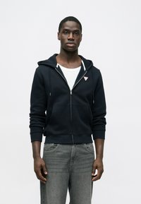 Námornícka mikina na zips s prednými vreckami, bielymi šnúrkami a nášivkou s logom. Nosená cez biele tričko, v kombinácii so sivými džínsami.