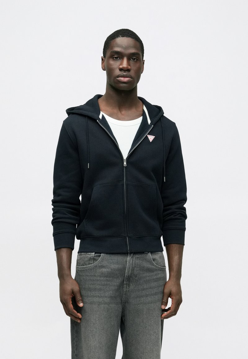 Námornícka mikina na zips s prednými vreckami, bielymi šnúrkami a nášivkou s logom. Nosená cez biele tričko, v kombinácii so sivými džínsami.