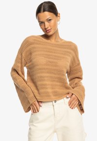Light brown knitted sweater featuring a loose fit, wide sleeves, and a textured striped pattern. Paired with cream-colored pants.