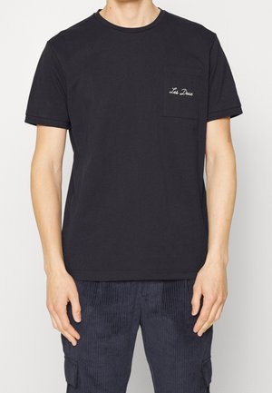 Camiseta de algodón negra con cuello redondo, mangas cortas y un bolsillo en el pecho izquierdo que presenta texto bordado en blanco. Textura suave.