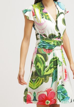 Mujer con vestido cruzado de manga corta con grandes hojas verdes y patrones florales en rosa brillante y rojo sobre un fondo blanco.