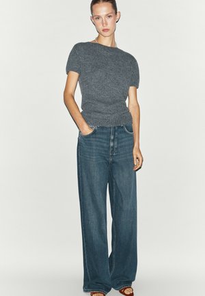 Frau trägt ein graues kurzärmeliges Strickoberteil und weit geschnittene blaue Jeans, steht mit einer Hand in der Tasche vor einem weißen Hintergrund.