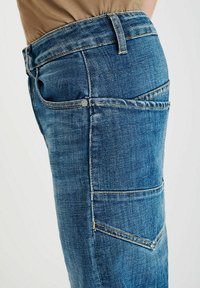 Close-up van blauwe spijkerbroek met zichtbare zijzak en riemlussen, gedragen met een beige top.