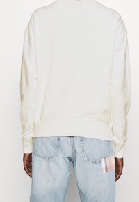 Světle krémová mikina s kulatým výstřihem a kleslými rameny, kombinovaná se světle modrými džíny, které mají na pravé kapse záplatu z kostkované látky.