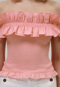 Off-the-shoulder rosa topp med volanghals og smocket stoff. Har en utsvingt kant og teksturert sømdetalj.