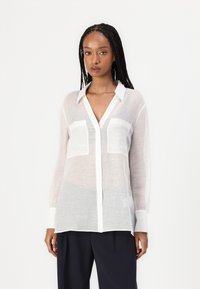 Camicia bianca trasparente a maniche lunghe con scollatura a V, design con bottoni e due tasche sul petto; presenta colletto e polsini bianchi a contrasto.