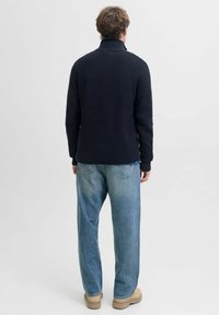 Suéter de punto azul marino oscuro con un cuello alto, patrón texturizado y dobladillo acanalado, combinado con pantalones vaqueros holgados de color azul claro y botas beige.