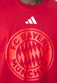 Red cotton T-shirt featuring a large, embossed FC Bayern München logo in the center and a white Adidas logo above.
