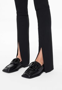 Mustat loafers, joissa on neliömäinen kärki, suurella pyöreällä soljella ja litteällä korolla. Yhdistetty tiukkojen mustien housujen kanssa, joissa on sivuslitit.