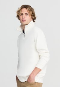 Pullover in pile bianco con zip fino al colletto, colletto alto, polsini a costine e vestibilità rilassata. Tessuto morbido con una finitura liscia.