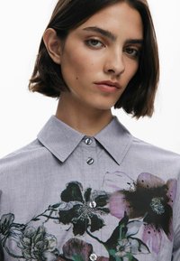 Camicia grigia con bottoni, decorata con un motivo floreale nei colori nero, viola e verde. Presenta un colletto e un tessuto liscio e texturizzato.