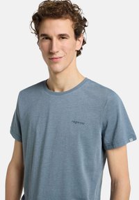 T-shirt en coton bleu à manches courtes, avec un logo discret sur la poitrine et une petite étiquette sur la manche. Texture lisse et coupe décontractée.