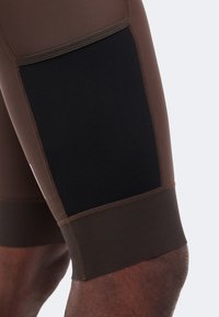 Καφέ αθλητικό σορτς με μεγάλο μαύρο δίχτυ στο μηρό, φορεμένα στο πόδι ενός ατόμου σε απλό φόντο.