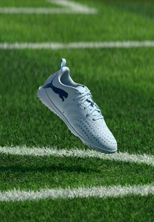 Ένα λευκό ποδοσφαιρικό παπούτσι Puma επιπλέει πάνω από πράσινο γρασίδι με λευκές γραμμές γηπέδου στο φόντο.