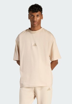Junger Mann steht, trägt beige Adidas T-Shirt und passende Hose mit Logo auf Brust und Oberschenkel, neutraler Hintergrund.