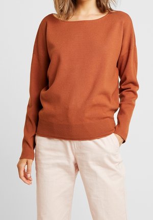 Femme portant un pull à manches longues couleur rouille et un pantalon beige clair, une main dans la poche, sur un fond uni.