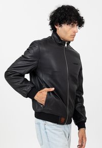 Giacca bomber in pelle nera con colletto alto, chiusura con zip, tasche laterali e polsini a coste. Presenta un'etichetta arancione del marchio all'orlo.