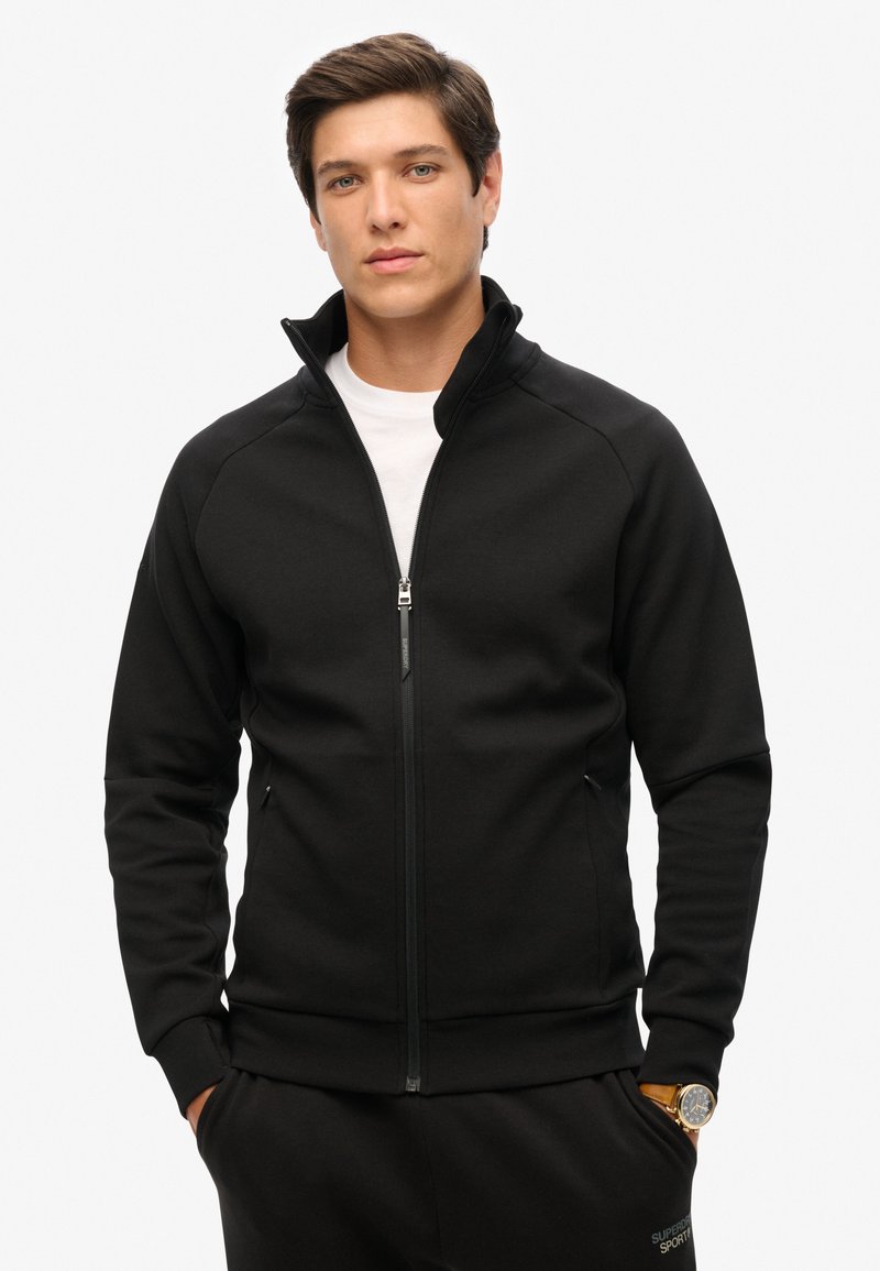 Schwarze Zip-Jacke aus weichem Stoff, mit hohem Kragen, Slim Fit und Frontreißverschluss. Kombiniert mit passenden dunklen Hosen und einer Uhr.