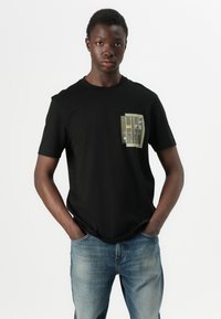 T-shirt nera a maniche corte con una toppa verde texturizzata caratterizzata da dettagli stampati, abbinata a jeans blu chiaro. Tessuto morbido, vestibilità casual.