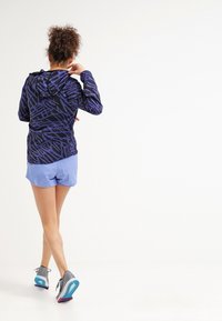 Chaqueta con capucha en azul marino con patrón abstracto en púrpura, combinada con pantalones cortos de color azul claro y zapatillas grises con acentos en azul verdoso y rosa.