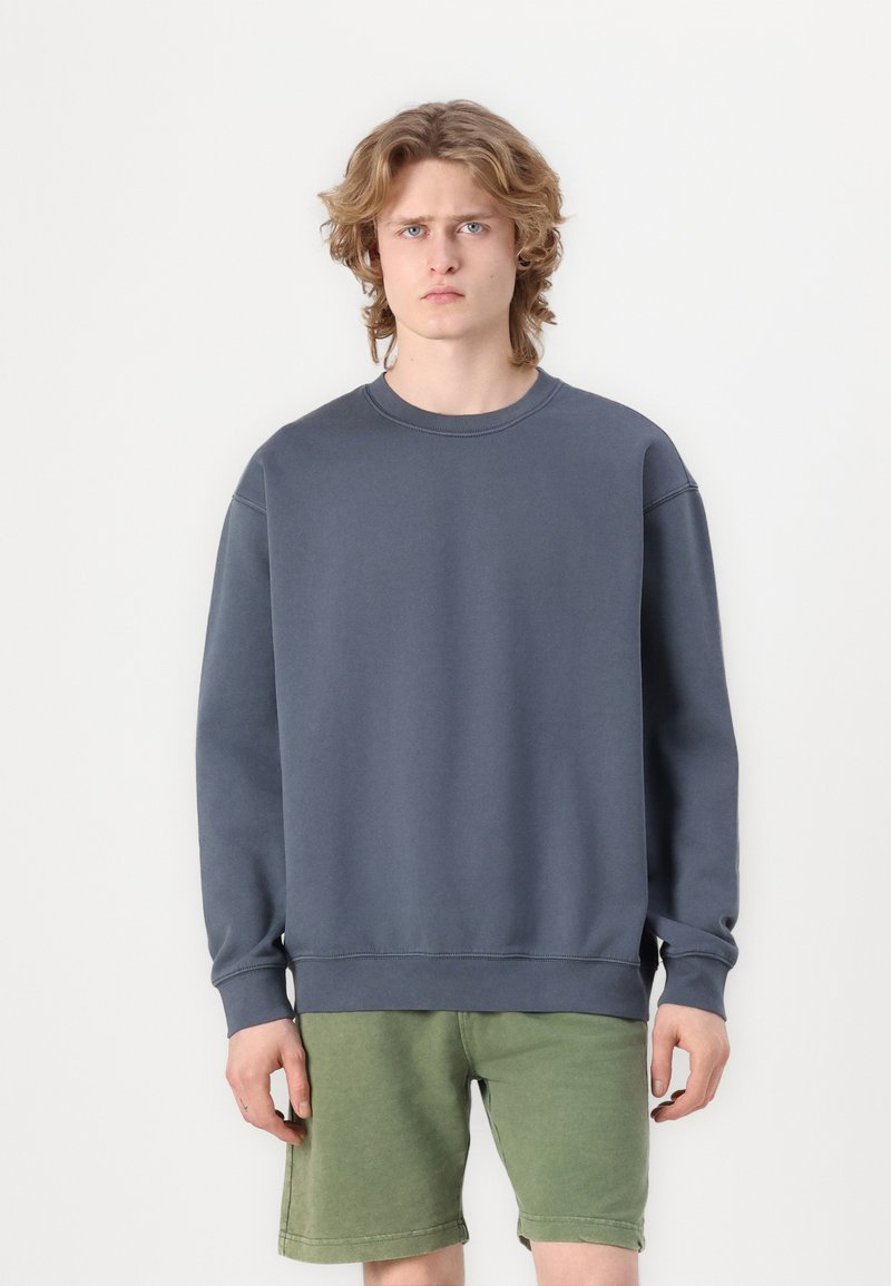 Sivý bavlnený crewneck sveter v zmesi bavlny, s rebrovanými manžetami a lemom. Nosí sa s zelenými šortkami, ktoré majú voľný strih a hladkú textúru.