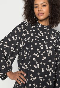 Blusa preta com padrão de margaridas brancas, decote alto, mangas bufantes com punhos franzidos, tecido leve e textura suave.