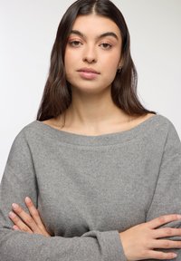 Maglione grigio oversize con scollo a barca, tessuto morbido e maniche lunghe. La modella posa con le braccia incrociate, mostrando il capo.