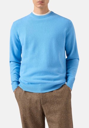 Uomo che indossa un maglione celeste girocollo e pantaloni marroni con trama, in piedi con le mani nelle tasche davanti a uno sfondo semplice.
