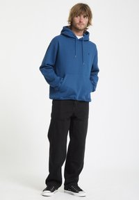 Blauwe hoodie met voorzak, capuchon met trekkoord en een logo. Gepaarde met zwarte wijde broek en zwarte sneakers. Effen witte achtergrond.
