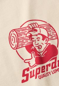 Beige t-shirt med en rød grafik af en smilende karakter, der holder en stamme, ledsaget af teksten "Superdry Quality Lumber" i fed skrift.