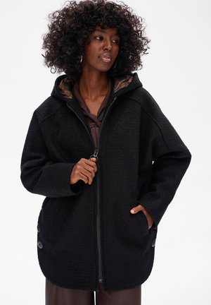 Zwarte fleece jas met ritssluiting, brede mouwen, een capuchon en decoratieve knopen aan de zijkanten. De binnenkant heeft een subtiele patroonvoering.