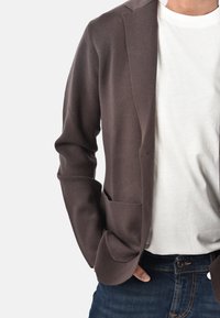 Uomo con blazer marrone sopra una t-shirt bianca e jeans blu, con le mani nelle tasche, su uno sfondo chiaro e uniforme.