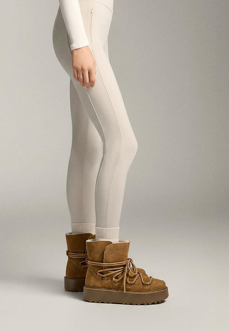 Cizme din suede maro cu o talpă chunky, având un design cu șireturi și căptușeală moale, asociate cu colanți ajustați de culoare deschisă.