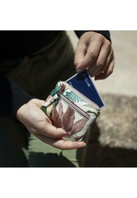 Cartera con patrón floral y cremallera, hecha de material suave, con hojas verdes y acentos rosas, sostenida en una mano, con una tarjeta parcialmente visible.
