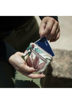 Cartera con patrón floral y cremallera, hecha de material suave, con hojas verdes y acentos rosas, sostenida en una mano, con una tarjeta parcialmente visible.