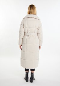 Kobieta w długim beżowym pikowanym płaszczu z paskiem w talii i czarnych botkach na obcasie, stojąca tyłem na białym tle.