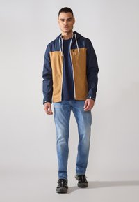 Vindjakke i navy og tan, med lynlås foran, justerbar hætte og snoretræk. Sammen med lyseblå jeans og sorte sneakers.