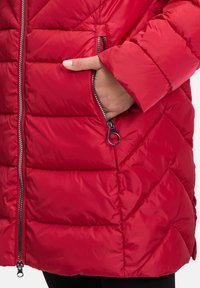 Detail einer roten Steppjacke mit diagonaler Steppung, mit Reißverschlusstaschen und einer glatten, glänzenden Stoffoberfläche.