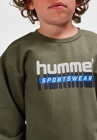 Olijfgroene sweatshirt met een ronde hals, voorzien van grote "hummel" en "SPORTSWEAR" teksten in witte en blauwe grafische letters. Zacht, textuurrijk materiaal.