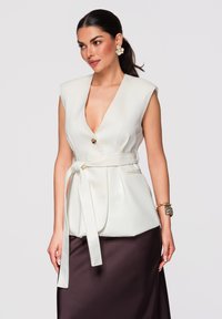 Femme portant un blazer blanc sans manches ceinturé et une jupe en satin foncé, avec de grandes boucles d’oreilles florales et des bracelets en or, debout devant un fond uni.